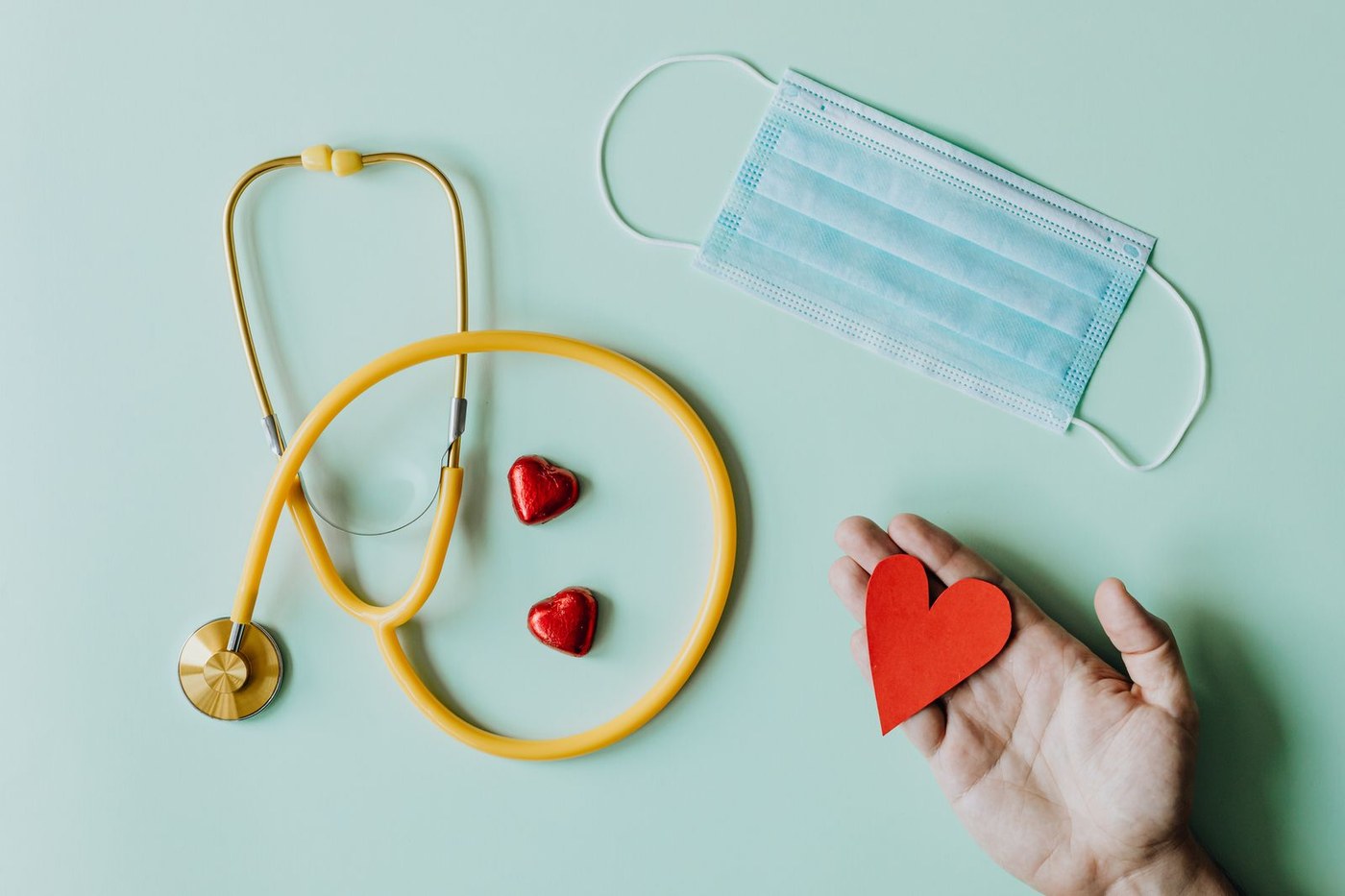 Yellow stethoscope coiled beside red heart shapes and a blue surgical mask on a light green background